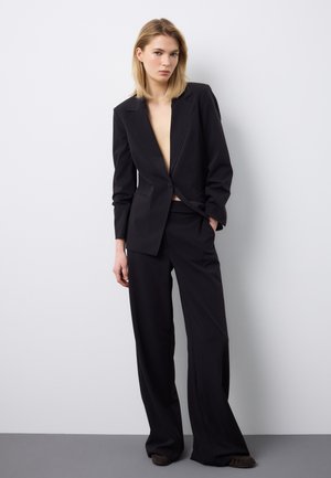 Mujer vistiendo un traje sastre negro con blazer de un solo botón, pantalones de pierna ancha, con la mano en el bolsillo, de pie contra una pared blanca lisa.