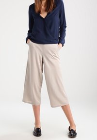 Top avvolgente blu navy con maniche lunghe abbinato a culotte beige a gamba larga. Scarpe nere slip-on completano l'outfit. Texture del tessuto liscia.