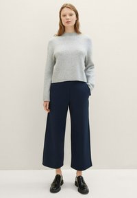 Ljust grå, ribbad tröja med hög halsringning, kombinerad med vida, marinblå byxor. Svarta snörskor fulländar outfiten.