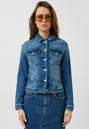 Frau trägt blaue, geknöpfte Jeansjacke und passenden Rock mit bernsteinfarben getönten, quadratischen Sonnenbrillen, steht vor hellem einfarbigem Hintergrund.