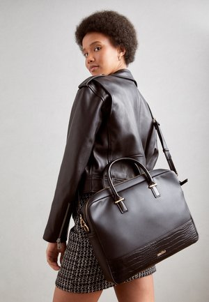 Jeune femme portant une veste en cuir noire et une jupe à motifs, tenant un grand sac à main en cuir noir sur l'épaule.