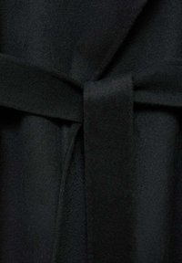 Manteau en laine noire avec une ceinture nouée à la taille montrant la texture et les plis du tissu.