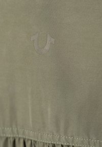 Un tissu vert olive au toucher lisse présente un logo proéminent dans une teinte plus claire. L'ourlet a un détail froncé, mettant en valeur le design.