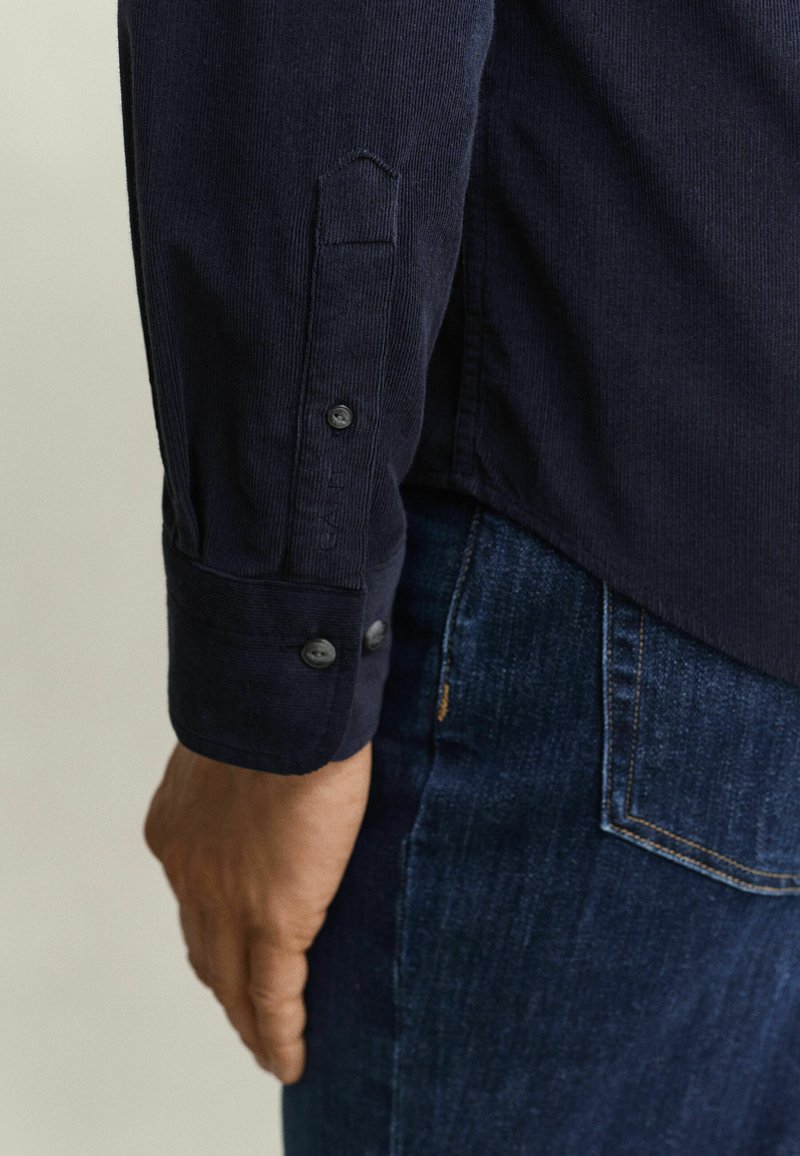 Man's left arm wearing a dark long-sleeve shirt with buttoned cuff next to blue denim jeans pocket.
