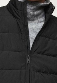 Veste sans manches noire matelassée avec un col montant, présentant un design matelassé horizontal et une fermeture éclair frontale. Portée par-dessus un pull à col roulé gris clair.