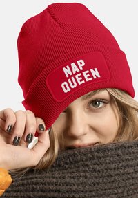 Red knit beanie featuring a rectangular patch with "NAP QUEEN" in bold white letters. Background shows a textured brown scarf.