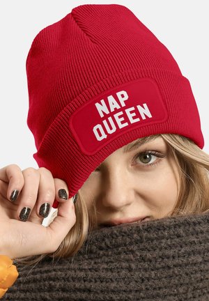 Red knit beanie featuring a rectangular patch with "NAP QUEEN" in bold white letters. Background shows a textured brown scarf.
