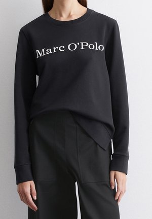 Schwarzes Sweatshirt mit langen Ärmeln und weißem Schriftzug „Marc O’Polo“, getragen mit schwarzen High-Waist-Hosen vor einfachem hellem Hintergrund.