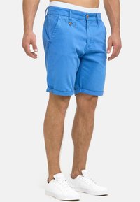 Homme portant un short bleu retroussé avec des boutons marron, des baskets blanches et une ceinture blanche, se tenant devant un fond blanc.