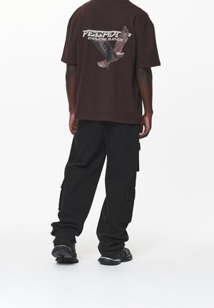 Persona vistiendo una camiseta de gran tamaño de color marrón oscuro con gráfico de águila y texto "Pegador Endless Silence", pantalones cargo negros y zapatillas negras, vista desde atrás.