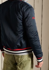 Giacca bomber color navy in tessuto lucido, con polsini a righe rosse e bianche, e un logo sulla spalla sinistra. Indossata sopra una maglietta grigia.