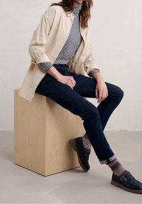 Beige button-up shirt over a striped turtleneck, dark blue jeans, patterned socks, and black shoes, seated on a wooden cube.