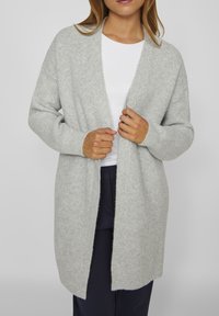 Světlý šedý cardigan s otevřeným předním dílem, žebrovanou texturou, dlouhými rukávy a volným střihem, nošený přes bílý top a tmavě modré kalhoty.