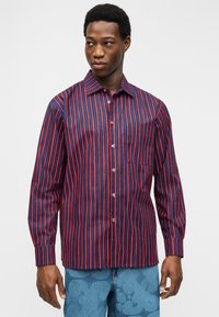 Άνδρας που φοράει πουκάμισο με μακριά μανίκια και κάθετες κόκκινες και μπλε ρίγες, στέκεται με τα χέρια κατεβασμένα, κοιτάζοντας προς τα αριστερά μπροστά σε απλό φόντο.