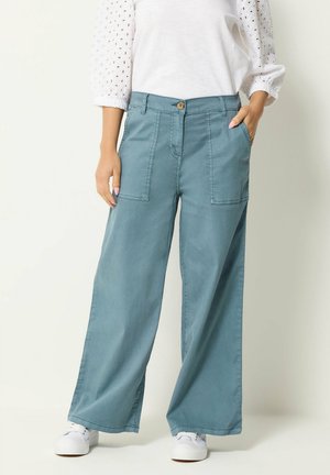 Donna che indossa pantaloni larghi verde acqua con tasche applicate frontali e sneakers bianche, in piedi con una mano in tasca.