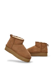Botas de tobillo de ante marrón con un suave forro de pelo sintético, punta redonda y una suela de goma texturizada baja. Presenta una lengüeta en la parte trasera.