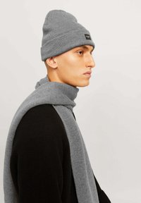 Beanie grigio lavorato a maglia con risvolto e etichetta discreta; sciarpa grigia abbinata si adagia su un maglione nero, evidenziando un motivo a maglia testurizzato.