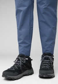 Scarpe da trekking nere con dettagli grigi, tessuto testurizzato e lacci riflettenti, abbinate a pantaloni da esterno blu. Il terreno è di un colore chiaro.