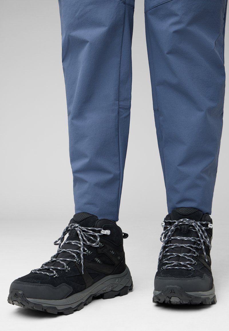 Scarpe da trekking nere con dettagli grigi, tessuto testurizzato e lacci riflettenti, abbinate a pantaloni da esterno blu. Il terreno è di un colore chiaro.