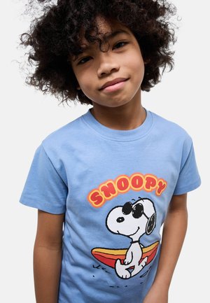Enfant aux cheveux bouclés portant un T-shirt bleu clair avec Snoopy assis sur une planche de surf et un texte rouge et jaune au-dessus.