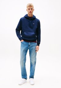 Νεαρός άνδρας που στέκεται, φορώντας μπλε ναυτικό φούτερ, ανοιχτό μπλε τζιν και λευκά αθλητικά παπούτσια, με το ένα χέρι στην τσέπη, μπροστά σε λευκό φόντο.