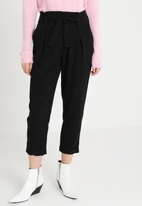 Pantaloni neri su misura con cintura a cravatta, caratterizzati da pieghe e lunghezza cropped. Abbinati a stivali alla caviglia bianchi e angolari.