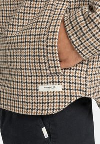 Brązowa, czarna i biała kurtka w wzór houndstooth z metką "ELEMENT CO." i widoczną kieszenią, zestawiona z ciemnymi spodniami.