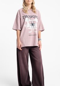 Przesadnie duży różowy t-shirt z nadrukiem graficznym przedstawiającym twarze i wzór gwiazdy. Noszony z luźnymi, ciemnofioletowymi spodniami.