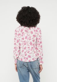 Donna con capelli ricci e scuri indossa una camicia bianca con motivo floreale rosa e jeans azzurri chiari, in piedi di spalle contro uno sfondo neutro.