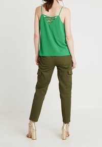 Top verde senza maniche con design incrociato sul retro, abbinato a pantaloni cargo verde oliva con tasche frontali e taglio alla caviglia, indossato con tacchi dorati.