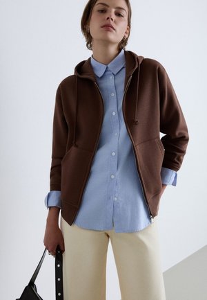 Jeune personne portant un sweat à capuche marron zippé sur une chemise bleu clair boutonnée et un pantalon crème, tenant un sac à main noir à ses côtés.