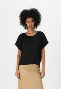 Maglietta nera a maniche corte con una vestibilità rilassata e scollo ampio, realizzata in un tessuto liscio. Indossata con una gonna a tubino beige liscia.