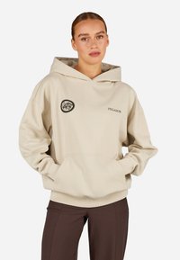 Béžová bavlnená mikina s predným vreckom, kapucňou na šnúrku a okrúhlym logom na ľavom prsníku. Uvoľnený strih a lemované manžety.