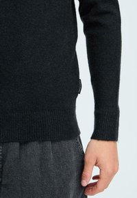 Schwarzer Strickpullover mit gerippten Bündchen, der eine weiche Textur aufweist. Seitliches Etikett sichtbar, kombiniert mit dunkelgrauen Hosen.