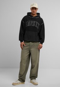 Svarte hettegenser med "MARKET" tekst, kengurulomme; olivengrønne løse bukser; kremfargede sneakers; beige caps med "Call My Lawyer" tekst.