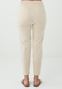 Een persoon staat met de rug naar de kijker, gekleed in lichtbeige broek en platte sandalen, met de armen ontspannen langs het lichaam tegen een effen achtergrond.