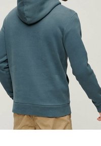 Hombre vestido con una sudadera con capucha azul y pantalones beige, visto desde atrás contra un fondo sencillo.