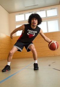 Melnā bezpiedurkņu krekliņš ar "Chicago Bulls" logo, melnas šortus ar baltām svītrām, melnas basketbola kurpes, driblējot basketbola bumbu uz sporta zāles grīdas.