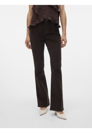 Mujer con pantalones de pana marrón oscuro de pierna recta, top marrón oscuro con volantes y tacones blancos de punta, de pie sobre un fondo blanco.