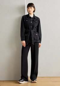 Giacca in denim nera con tasche frontali e cintura, abbinata a pantaloni neri a gamba larga. Modella con sneaker scure, in piedi su un pavimento in legno.