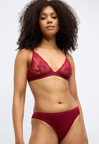 Burgundowa koronkowa trójkątna bielizna i pasujące figi z cienkimi ramiączkami. Kwiatowy wzór na biustonoszu; gładki materiał na figach. Podkreślona dokładność dopasowania.