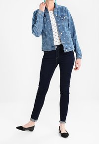 Ljusblå denimjacka med hjärtan, bärs över en vit skjorta med små mönster, i kombination med mörkblå skinny jeans och svarta ballerinas.