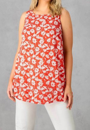 Frau trägt ärmelloses rotes Oberteil mit weißem Blumenmuster und weiße Hose, steht vor grauem Hintergrund.