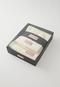 Set di cappello e sciarpa lavorati a maglia in crema, confezionato in una scatola nera. Presenta una texture a coste e etichette con accenti rossi con il marchio "HUNTER".