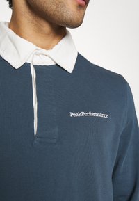 Marineblauwe katoenen polo met een witte corduroy kraag, voorzien van een knoopsluiting en het "PeakPerformance"-logo in witte borduursel.