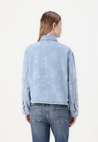 Veste en jean bleu clair avec un motif floral, dotée d'un col et de manches longues, complétée par une coupe décontractée et des poches latérales.