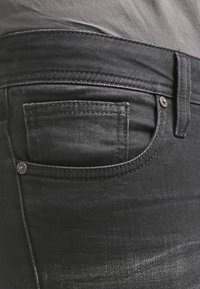 Černé džíny z denimu s úzkým střihem, s kapsou na zádech zdobenou šitím a kovovými nýty na pásku.