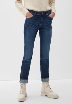 Jean en denim bleu foncé avec une coupe slim, présentant des ourlets roulés et cinq poches. Portés avec des bottes à lacets de couleur crème.