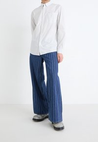 Camicia a maniche lunghe a righe bianche con taschino frontale, abbinata a pantaloni in denim blu a gamba larga con righe verticali bianche e stivali grigi.