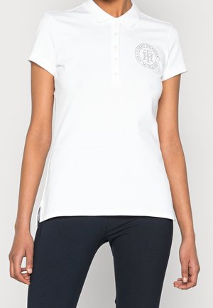 Vrouw in een wit poloshirt met korte mouwen, knopen en een rond logo op de linkerkant van de borst, gecombineerd met zwarte broek.